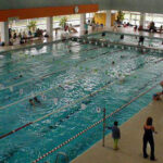Hallenbad mit mehreren Personen, die in Bahnen schwimmen; andere sitzen oder stehen um den Pool herum. Große Fenster sorgen für natürliches Licht.