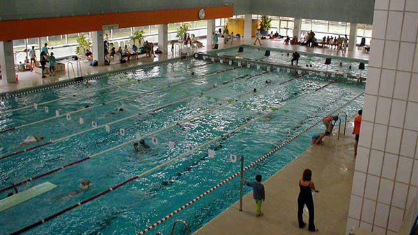 Hallenbad mit mehreren Personen, die in Bahnen schwimmen; andere sitzen oder stehen um den Pool herum. Große Fenster sorgen für natürliches Licht.