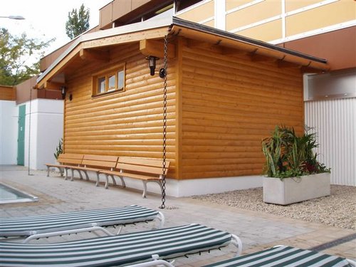 Eine kleine Holzhütte mit schrägem Dach steht neben Bänken, Topfpflanzen und gestreiften Liegestühlen in der Nähe eines Swimmingpools.