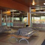 Innenwartebereich mit Metallbänken vor großen Fenstern mit Blick auf ein Schwimmbad. Der Bereich hat geflieste Böden und eine Holzdecke mit Oberlichtern.