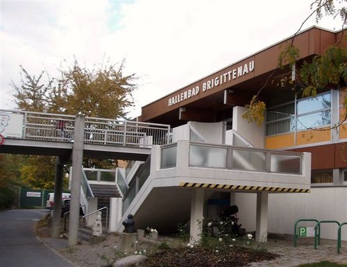 Außenansicht des Ballenbads Brigittenau, eines öffentlichen Gebäudes mit einer Treppe, die zu einem Eingang führt, und großen Fenstern, umgeben von Bäumen und einem gepflasterten Gehweg.