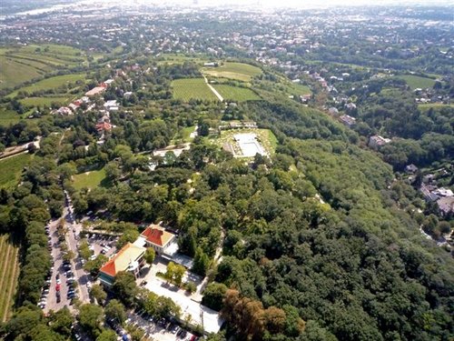 Luftaufnahme einer grünen, hügeligen Landschaft mit verstreuten Gebäuden, Parkplätzen und Bäumen, die sich unter klarem Himmel in Richtung einer entfernten Stadtlandschaft erstreckt.