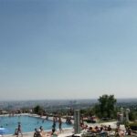 Die Menschen entspannen sich und nehmen ein Sonnenbad an einem auf einem Hügel gelegenen Freibad, während im Hintergrund eine Stadtlandschaft unter einem klaren Himmel zu sehen ist.