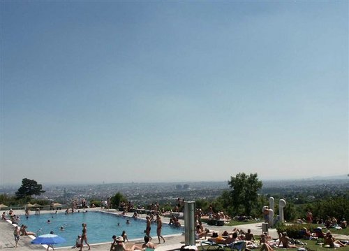 Die Menschen entspannen sich und nehmen ein Sonnenbad an einem auf einem Hügel gelegenen Freibad, während im Hintergrund eine Stadtlandschaft unter einem klaren Himmel zu sehen ist.