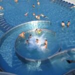 Luftaufnahme eines überfüllten Freibads mit schwimmenden, watenden und spielenden Menschen in einem kreisförmigen Beckenbereich, der mit dem Hauptbecken verbunden ist.