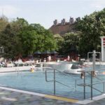 Öffentliches Freibad mit schwimmenden und faulenzenden Menschen, umgeben von Bäumen, Spielgeräten und nahe gelegenen Gebäuden.