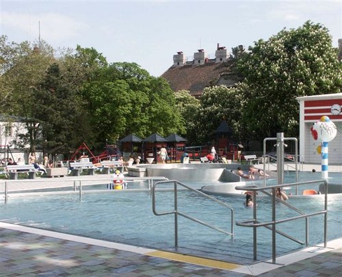 Öffentliches Freibad mit schwimmenden und faulenzenden Menschen, umgeben von Bäumen, Spielgeräten und nahe gelegenen Gebäuden.