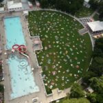 Luftaufnahme eines Außenschwimmbads mit Menschen im Wasser und einer großen Rasenfläche in der Nähe, auf der sich Menschen auf Handtüchern und Stühlen sonnen.