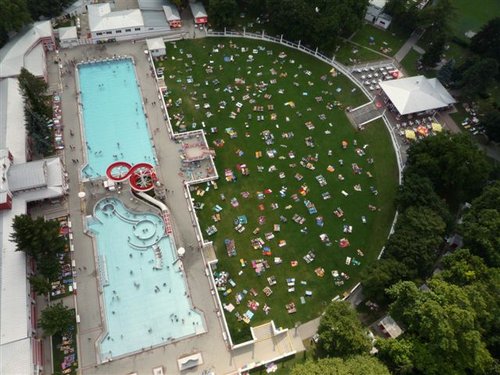 Luftaufnahme eines Außenschwimmbads mit Menschen im Wasser und einer großen Rasenfläche in der Nähe, auf der sich Menschen auf Handtüchern und Stühlen sonnen.