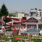 Die Menschen entspannen sich auf Liegestühlen und im Gras in der Nähe eines Außenpools mit einer rot-weißen Spiralwasserrutsche, umgeben von Gebäuden und Bäumen.