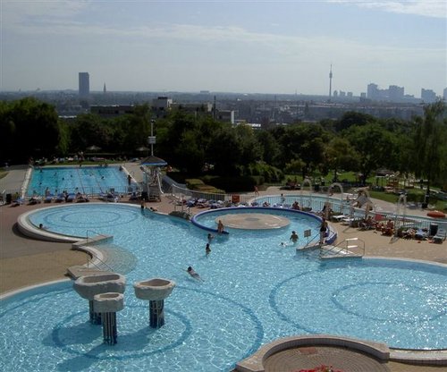 Öffentlicher Freibadkomplex mit mehreren Becken, schwimmenden und faulenzenden Menschen und einer im Hintergrund sichtbaren Stadtsilhouette bei teilweise bewölktem Himmel.