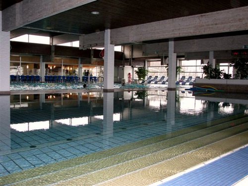 Hallenbad mit klarem Wasser, gefliestem Boden, Liegestühlen an der Seite und großen Fenstern, die viel Tageslicht hereinlassen.