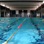 Hallenbad mit Bahnen, die durch ein schwimmendes Seil getrennt sind, ein paar Schwimmer im Wasser und Liegestühle an den Wänden.