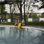 Ein kleines Außenbecken mit einer gelben Rutsche zwischen zwei Tierfiguren, umgeben von Bäumen und blau-weiß gestreiften Gebäuden im Hintergrund.