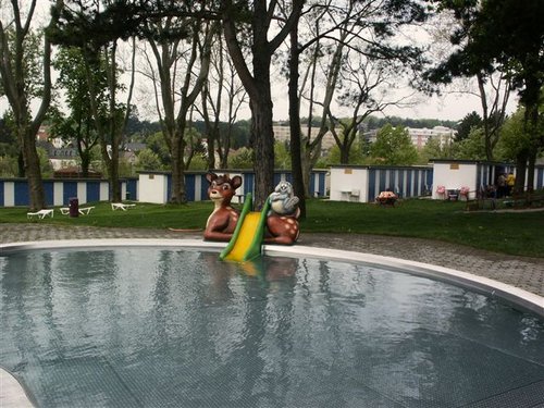 Ein kleines Außenbecken mit einer gelben Rutsche zwischen zwei Tierfiguren, umgeben von Bäumen und blau-weiß gestreiften Gebäuden im Hintergrund.