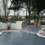 Ein Kinderbecken im Freien mit Tierfiguren, einer kleinen Rutsche und umliegenden Bäumen; das Wasser scheint ruhig und der Himmel ist bedeckt.