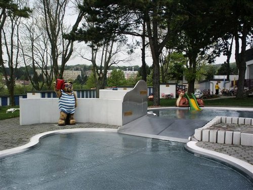 Ein Kinderbecken im Freien mit Tierfiguren, einer kleinen Rutsche und umliegenden Bäumen; das Wasser scheint ruhig und der Himmel ist bedeckt.
