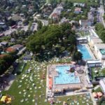 Luftaufnahme eines öffentlichen Freibadkomplexes mit Menschen, die sich auf der Wiese sonnen, umgeben von Bäumen und Wohngebäuden.