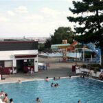 Außenschwimmbad mit Menschen im Wasser und in der Nähe; im Hintergrund sind ein Gebäude, Bäume und bunte Wasserrutschen zu sehen.