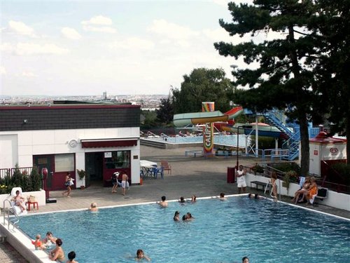 Außenschwimmbad mit Menschen im Wasser und in der Nähe; im Hintergrund sind ein Gebäude, Bäume und bunte Wasserrutschen zu sehen.