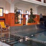 Hallenbad mit Wasserrutsche, Metallgeländer, gelbe Liegestühle, orangefarbene Kacheln, Topfpflanzen und große Fenster, die für Tageslicht sorgen.