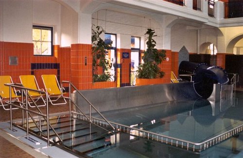 Hallenbad mit Wasserrutsche, Metallgeländer, gelbe Liegestühle, orangefarbene Kacheln, Topfpflanzen und große Fenster, die für Tageslicht sorgen.