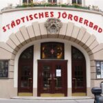 Eingang des Städtischen Jörgerbads mit Rundbogenportal, Glasscheiben und roter Beschilderung über der Tür.