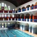 Hallenbad mit hohen Bogenfenstern, zwei Ebenen mit blauen Balkonen, gefliesten Wänden und mehreren Schwimmern im Becken.