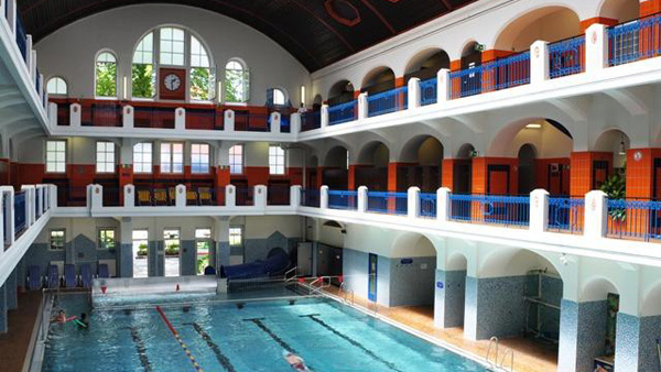 Hallenbad mit hohen Bogenfenstern, zwei Ebenen mit blauen Balkonen, gefliesten Wänden und mehreren Schwimmern im Becken.