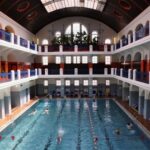 Hallenbad in einem großen, historischen Gebäude mit Bogenfenstern, mehreren Balkonen und Schwimmern auf bestimmten Bahnen.