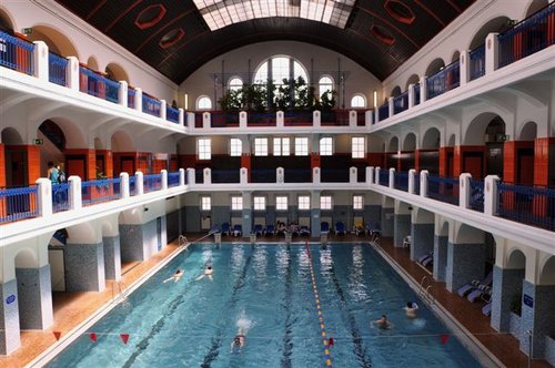 Hallenbad in einem großen, historischen Gebäude mit Bogenfenstern, mehreren Balkonen und Schwimmern auf bestimmten Bahnen.