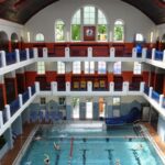 Hallenbad mit drei Ebenen, großen Bogenfenstern, Uhr an der hinteren Wand und mehreren Personen, die im Becken schwimmen.