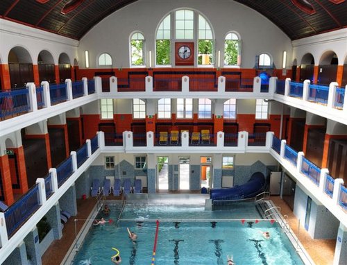 Hallenbad mit drei Ebenen, großen Bogenfenstern, Uhr an der hinteren Wand und mehreren Personen, die im Becken schwimmen.