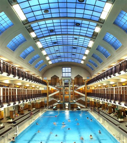 Hallenbad mit Personenschwimmen, umgeben von zwei Ebenen mit Spinden und Bänken und einer großen gewölbten Glasdecke, die natürliches Licht hereinlässt.