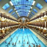 Hallenbad unter einem großen Glasdach, in dem Menschen schwimmen und sich entspannen; Umkleideräume und Balkone säumen beide Seiten des Schwimmbereichs.