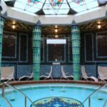 Innenpool mit Mosaikfliesen, umgeben von grünen Säulen und weißen Liegestühlen, unter einer Kuppel aus Buntglas.