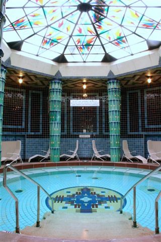 Innenpool mit Mosaikfliesen, umgeben von grünen Säulen und weißen Liegestühlen, unter einer Kuppel aus Buntglas.