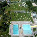 Luftaufnahme eines Freibadkomplexes mit zwei Becken, einer gelben Wasserrutsche und Grünflächen, auf denen sich die Menschen sonnen und entspannen.