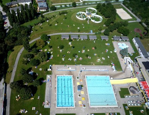 Luftaufnahme eines Freibadkomplexes mit zwei Becken, einer gelben Wasserrutsche und Grünflächen, auf denen sich die Menschen sonnen und entspannen.