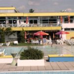 Swimmingpool im Freien mit verstreuten Liegestühlen, Tischen und Sonnenschirmen und einem modernen gelb-weißen Gebäude im Hintergrund.