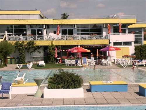 Swimmingpool im Freien mit verstreuten Liegestühlen, Tischen und Sonnenschirmen und einem modernen gelb-weißen Gebäude im Hintergrund.