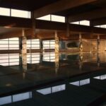 Hallenbad mit ruhigem Wasser, umgeben von großen Fenstern und Betonsäulen, in einer schwach beleuchteten, leeren Anlage.