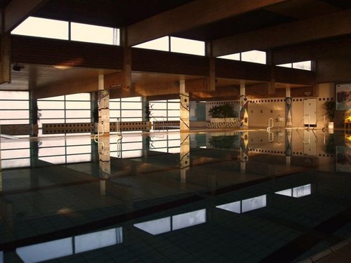 Hallenbad mit ruhigem Wasser, umgeben von großen Fenstern und Betonsäulen, in einer schwach beleuchteten, leeren Anlage.