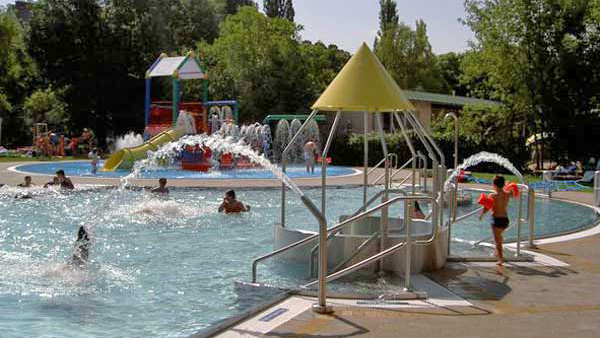 Kinder und Erwachsene spielen im Freibad mit Wasserfontänen, Rutschen und bunten Spielgeräten, die von Bäumen umgeben sind.