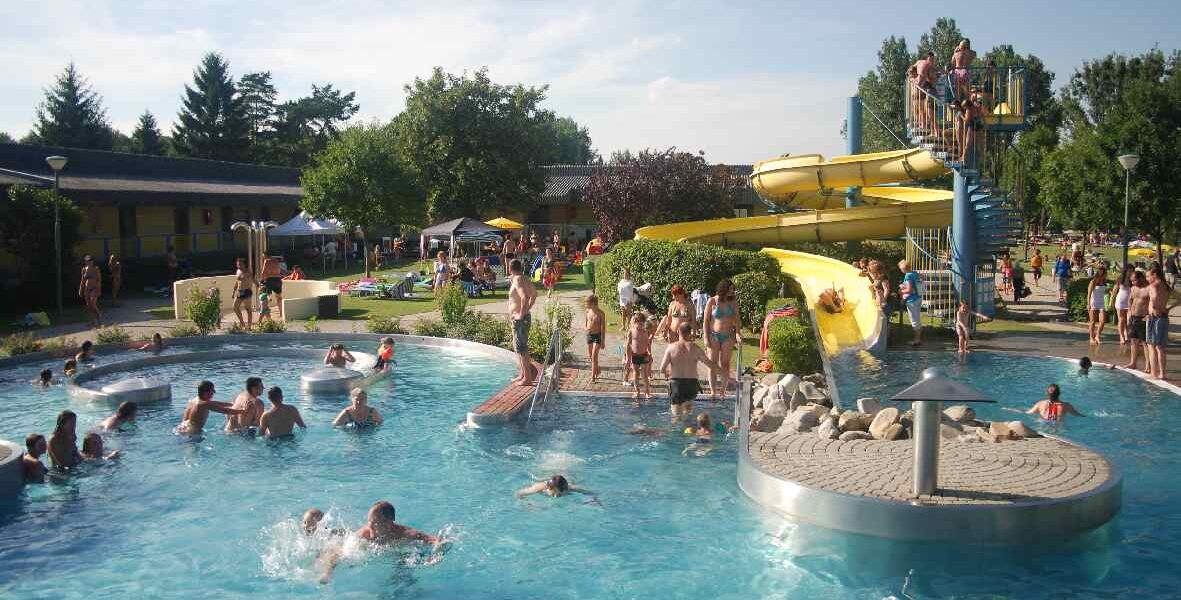 Öffentliches Freibad mit Menschen, die an einem sonnigen Tag schwimmen, faulenzen und darauf warten, die gelben Wasserrutschen zu benutzen. Bäume und Rasenflächen umgeben den Poolbereich.