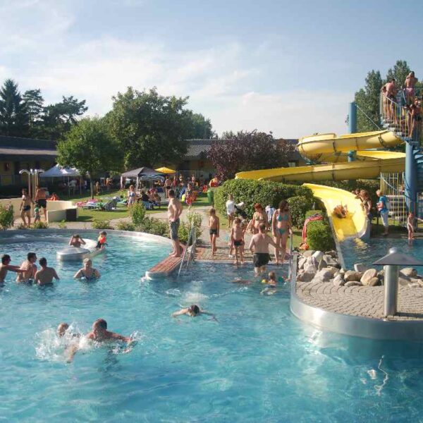 Öffentliches Freibad mit Menschen, die an einem sonnigen Tag schwimmen, faulenzen und darauf warten, die gelben Wasserrutschen zu benutzen. Bäume und Rasenflächen umgeben den Poolbereich.