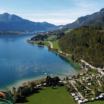 Eine Frau in traditioneller Kleidung blickt auf einen See, umgeben von grünen Hügeln, Bergen und einer kleinen Stadt unter einem klaren blauen Himmel.