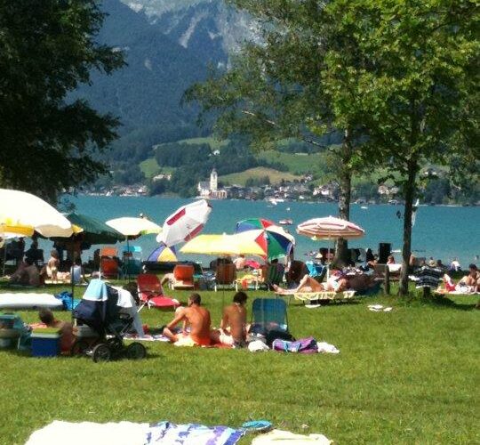 Menschen, die sich auf einer Wiese an einem See sonnen und entspannen, mit Sonnenschirmen und Handtüchern; im Hintergrund sind Berge und eine Kirche zu sehen.