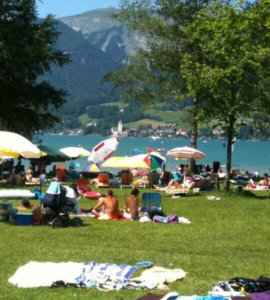 Menschen, die sich auf einer Wiese an einem See sonnen und entspannen, mit Sonnenschirmen und Handtüchern; im Hintergrund sind Berge und eine Kirche zu sehen.