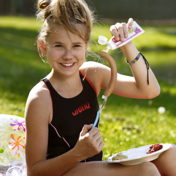 Ein junges Mädchen in einem schwarzen Badeanzug sitzt im Freien und drückt lächelnd Mayonnaise auf ein Würstchen, das auf einer Gabel über einem Teller liegt.